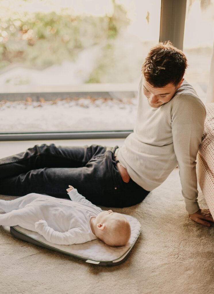 Vater sitzt am Boden und schaut auf das Kind, was neben ihm liegt. Beitragsbild zum Thema Familienfreundlichkeit im Unternehmen