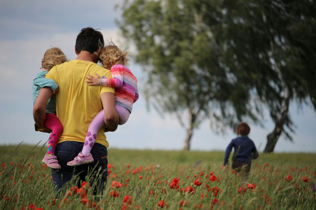 Vater von hinten zu sehen mit zwei Kindern auf dem Arm auf einer Wiese. Ein Kind läuft vorne weg. Beitragsbild zum Thema Familienfreundlichkeit im Unternehmen